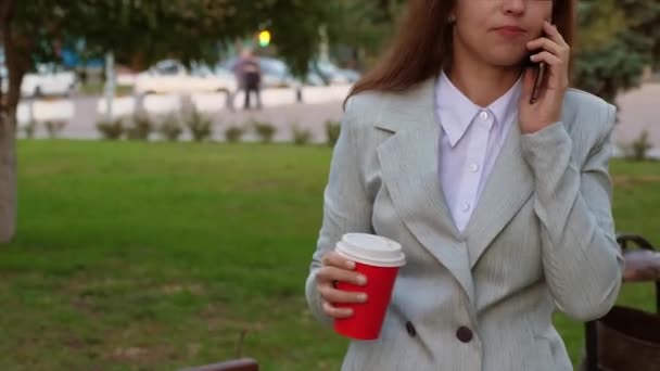 Mujer de negocios con gafas en traje ligero hablando en smartphone y tomando café en el parque — Vídeos de Stock