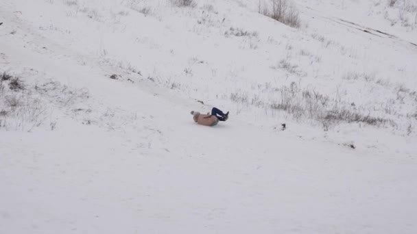 Jenta ruller ned bakken i slede i snø. Hun leker om vinteren i parken. Vinterleker. Kalde klimatiske forhold. Snødekte fjell for ski. Julehøytider – stockvideo