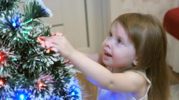 Feliz bebé está jugando cerca del árbol de Navidad y sonriendo. La chica acaricia su hermosa pícea peluda. Abeto inteligente. Año Nuevo. Vacaciones de Navidad — Vídeo de stock