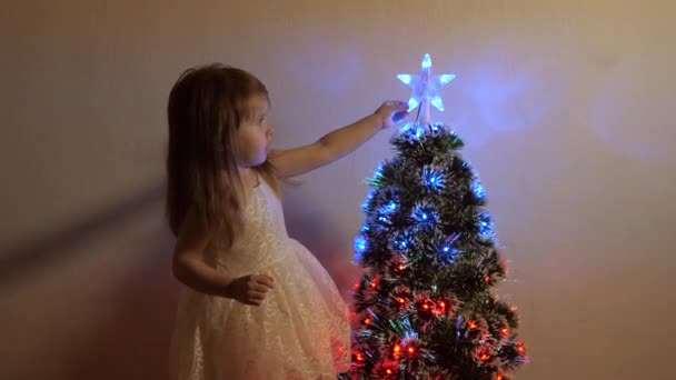 Çocuklar için mutlu noeller tatili konsepti. Çocuk, Noel ağacındaki bir Noel yıldızını inceliyor. Küçük kız, çocuk odasındaki bir Noel ağacının yanında oynuyor. Güzel yapay Noel ağacı. — Stok video