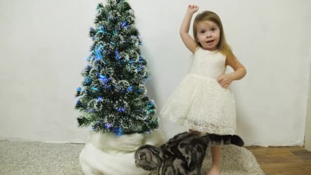Egy kislány játszik egy karácsonyfa mellett egy gyerekszobában egy macskával. Baba és karácsonyfa. Karácsonyi ünnepek — Stock videók