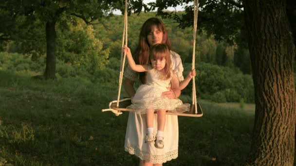 Concepto de una familia feliz. Mamá sacude a su hija en columpio bajo un árbol bajo el sol. madre y el bebé montar en columpio cuerda en una rama de roble en el bosque. Diversión familiar en el parque, en la naturaleza . — Vídeo de stock