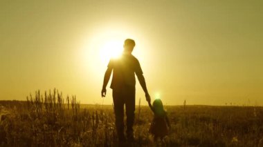Küçük bir çocuk ve baba, güzel bir gün batımının ışınlarında el ele tutuşarak tarlada yürür. Mutlu aile kavramı, mutlu çocukluk kavramı. Küçük kız babasının elini tutuyor, mutlu aile çayırda yürüyor..