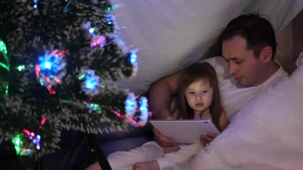 Padre e hija en la noche de Navidad, jugar y ver dibujos animados en la tableta, en una habitación para niños en una tienda de campaña con guirnaldas. bebé y papá están jugando en la habitación. concepto de infancia feliz y familia . — Vídeos de Stock