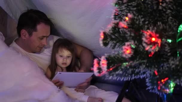 Noel akşamında baba ve kızı, çelenklerle dolu bir çadırda, tablet üzerinde çizgi film izleyip oynuyorlar. Bebek ve babam odada oynuyorlar. Mutlu çocukluk ve aile kavramı. — Stok video