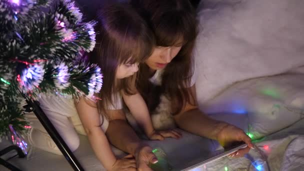 Μαμά και κόρη το βράδυ των Χριστουγέννων, παίζουν και να παρακολουθήσουν κινούμενα σχέδια στο tablet, σε ένα παιδικό δωμάτιο σε μια σκηνή με γιρλάντες. Το μωρό και η μητέρα παίζουν στο δωμάτιο. έννοια της ευτυχισμένης παιδικής ηλικίας και της οικογένειας. — Αρχείο Βίντεο