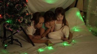 Aile Noel 'i konsepti. Noel akşamında çocuklar tabletle, çocuk odasında kanepede, çelenklerle oynarlar. Kız kardeşler çok renkli bir yılbaşı ağacıyla süslenmiş bir odada oynarlar.
