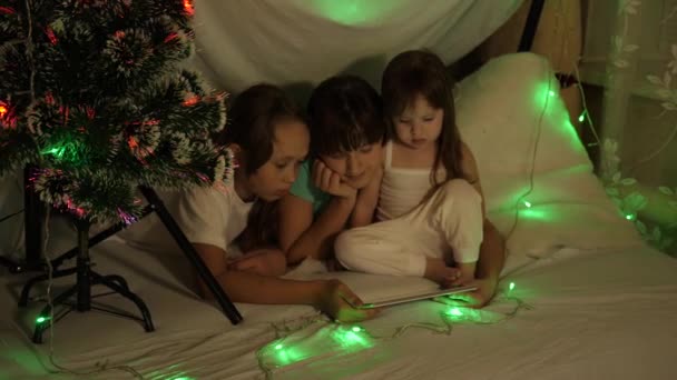 Concepto de una Navidad familiar. niños en el juego de la noche de Navidad en la tableta, en la habitación de los niños en el sofá, en la tienda con guirnaldas. hermanas juegan en una habitación decorada con un árbol de Año Nuevo multicolor — Vídeo de stock