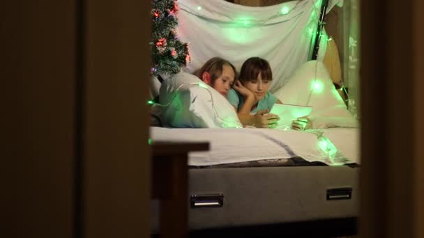 Діти грають у кімнаті, прикрашеній різнокольоровою новорічною ялинкою. сестри на різдвяний вечір грають на планшеті, в дитячій кімнаті на дивані, в наметі з гірляндами. концепція сім'ї Різдво . — стокове відео