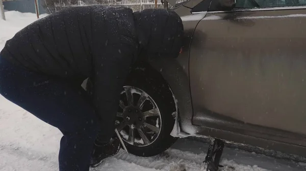 man raises car jack on road. man is changing wheel of car. replacement wheels in winter on road in a blizzard and snowfall. Breakdown of car, wheel change.