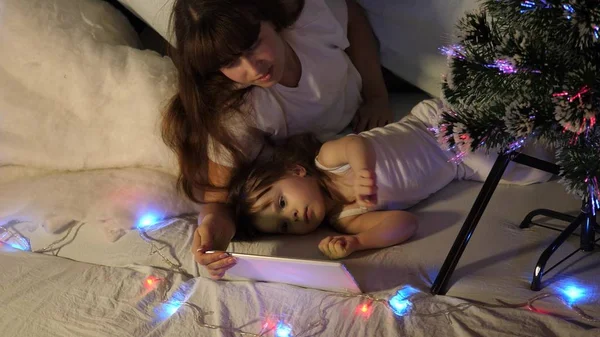happy mother and daughter on Christmas evening, play cartoons and watch cartoons on tablet, in a childrens room in a tent with garlands. baby and mother are playing in room. happy childhood concept.