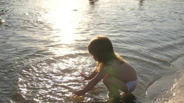 O bebé a espirrar na água. a salpicar água. Criança bonito brinca alegremente na praia. criança feliz banha-se no pôr-do-sol em um rio, mar, lago. conceito de infância feliz . — Fotografia de Stock