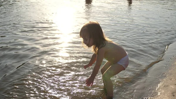 O bebé a espirrar na água. a salpicar água. Criança bonito brinca alegremente na praia. criança feliz banha-se no pôr-do-sol em um rio, mar, lago. conceito de infância feliz . — Fotografia de Stock