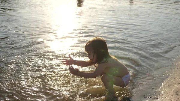 Baby planscht im Wasser. Spritzwasser. Nettes Kind spielt fröhlich am Strand. glückliches Kind badet im Sonnenuntergang auf einem Fluss, Meer, See. Konzept der glücklichen Kindheit. — Stockfoto