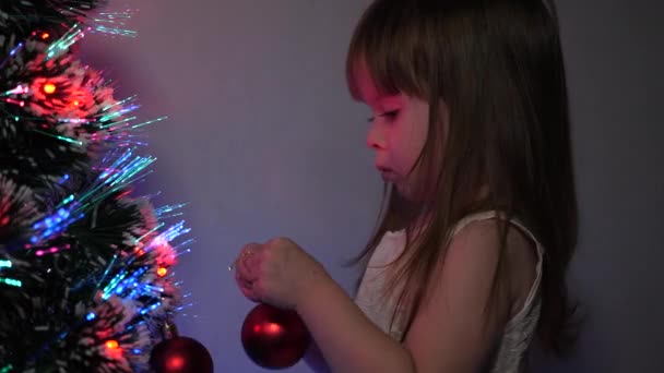 Kind versiert de kerstboom met kerstballen. close-up. klein kind speelt bij de kerstboom in de kinderkamer. dochter onderzoekt bloemenslinger op kerstboom. gelukkige kindertijd concept. — Stockvideo