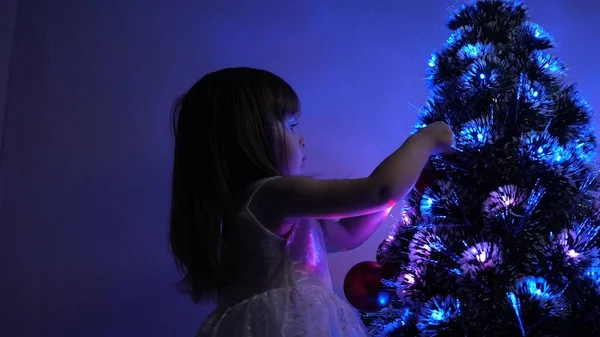 Pequena criança joga pela árvore de Natal no quarto das crianças. criança decora a árvore de Natal com bolas de Natal. filha examina guirlanda na árvore de Natal. conceito de infância feliz . — Fotografia de Stock