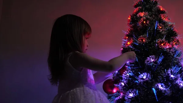 Kind versiert de kerstboom met kerstballen. klein kind speelt bij de kerstboom in de kinderkamer. dochter onderzoekt bloemenslinger op kerstboom. gelukkige kindertijd concept. — Stockfoto
