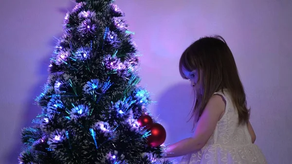 小孩用圣诞球装饰圣诞树。 小孩在儿童房的圣诞树旁玩耍。 女儿在圣诞树上检查花环。 快乐童年的概念. — 图库照片