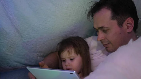 在圣诞节的晚上，父亲和女儿在一个有花环的帐篷里的儿童房间里，在平板电脑上玩和看卡通片。 爸爸和宝宝在房间里玩耍。 幸福童年和家庭的概念. — 图库照片