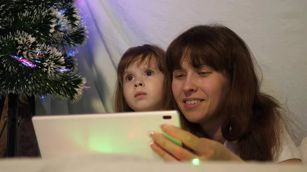 圣诞节的晚上，妈妈和女儿在一个有花环的帐篷里的儿童房间里，在平板电脑上玩和看卡通片。 婴儿和母亲正在房间里玩耍。 幸福童年和家庭的概念. — 图库照片