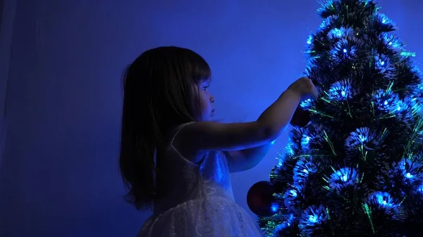 Pequena criança joga pela árvore de Natal no quarto das crianças. criança decora a árvore de Natal com bolas de Natal. filha examina guirlanda na árvore de Natal. conceito de infância feliz . — Fotografia de Stock