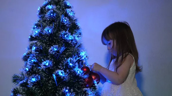 Kind versiert de kerstboom met kerstballen. klein kind speelt bij de kerstboom in de kinderkamer. dochter onderzoekt bloemenslinger op kerstboom. gelukkige kindertijd concept. — Stockfoto