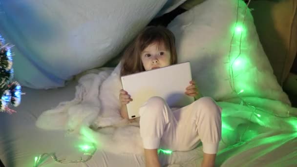 子供部屋のソファの上にタブレットコンピュータを持つかわいい女の子。子供部屋でクリスマスの夜にタブレットを持つ赤ちゃんの男の子。子供はPDA上の漫画を見る — ストック動画