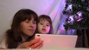 Çocuk ve anne odada tablet bilgisayarla oynuyorlar. Mutlu bir çocukluk ve aile kavramı. Anne ve kızı Noel akşamı çelenklerle dolu bir odada tabletten çizgi film izliyor..