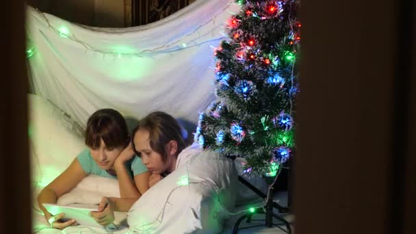 Koncepcja rodzinnych świąt. dzieci na Boże Narodzenie bawią się na tablecie, w pokoju dziecięcym na podwiązkach kanapy. siostry bawią się w pokoju ozdobionym wielokolorowym drzewem noworocznym — Wideo stockowe