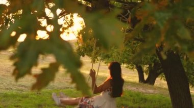 çocuk park günbatımında bir meşe dalı üzerinde bir ip salıncak sürmek. Close. Kız güler, sevinir. genç kız güneşte bir ağacın altında salıncakta sallanan, çocuklarla oynarken. Doğada aile eğlencesi. Yavaş çekim