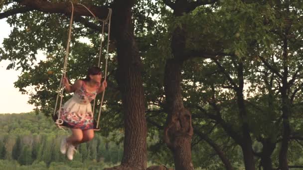 Niño balanceándose en un columpio en el parque bajo el sol. joven chica balanceándose en cuerda columpio en una rama de roble. adolescente chica disfruta de vuelo en swing en verano por la noche en el bosque. concepto de familia feliz y la infancia. — Vídeo de stock