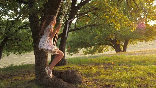 Barn som svänger på en gunga i parken i solen. ung flicka svänger på rep gunga på en ek gren. tonåring flicka njuter av flygning på swing på sommaren kväll i skogen. begreppet lycklig familj och barndom. — Stockfoto