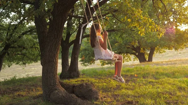Barn som svänger på en gunga i parken i solen. ung flicka svänger på rep gunga på en ek gren. tonåring flicka njuter av flygning på swing på sommaren kväll i skogen. begreppet lycklig familj och barndom. — Stockfoto