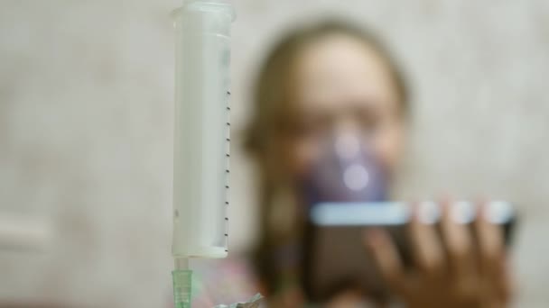 Egy maszkos beteg lány belélegez egy spray palackot, és számítógépes játékot játszik egy táblagépen. A kék kórházi maszkos kölyök belélegzi a gőzt a légutaktól a tüdőbe. Köhögés elleni kezelés. Közelkép — Stock videók