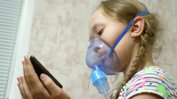 Una chica enferma en una máscara inhala una botella de spray y juega un juego de ordenador en una tableta. El niño con una máscara azul del hospital inhala humos desde las vías respiratorias hasta los pulmones. Tratamiento para la tos. Primer plano — Vídeo de stock