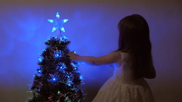 Dziecko bada gwiazdkę na drzewie świątecznym. Mała dziewczynka bawi się blisko choinki w pokoju dziecięcym. Piękna sztuczna choinka. Koncepcja szczęśliwego dzieciństwa. — Zdjęcie stockowe