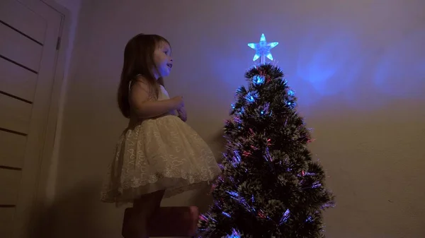 Feliz Navidad concepto de vacaciones para los niños. niño examina una estrella de Navidad en un árbol de vacaciones. La niña juega cerca de un árbol de Navidad en una habitación para niños. hermoso árbol de Navidad artificial . —  Fotos de Stock