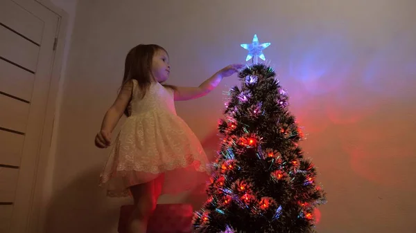 Conceito de férias de Natal feliz para crianças. a criança examina uma estrela de Natal em uma árvore de férias. menina brinca perto de uma árvore de Natal em uma sala de crianças. bela árvore de Natal artificial . — Fotografia de Stock