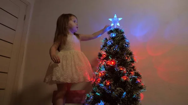 Conceito de férias de Natal feliz para crianças. a criança examina uma estrela de Natal em uma árvore de férias. menina brinca perto de uma árvore de Natal em uma sala de crianças. bela árvore de Natal artificial . — Fotografia de Stock