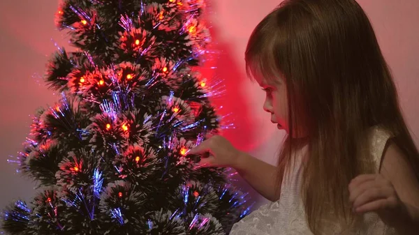 Filha examina guirlanda na árvore de Natal. pequena criança joga pela árvore de Natal no quarto das crianças. bela árvore de Natal artificial. conceito de infância feliz. Família joga para feriados de Natal — Fotografia de Stock