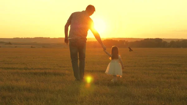 Figlioletta cammina con il padre in un prato tenendosi per mano. Il bambino tiene per mano i padri. famiglia cammina in serata fuori città. Papa 'e il bambino stanno riposando nel parco. bambino gioca con suo padre — Foto Stock