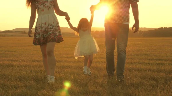 Liten dotter hoppar hålla händerna på pappa och mamma i parken på bakgrund av solen. Familjekoncept. barn leker med pappa och mamma på fältet i solnedgången ljus. Promenader med en liten unge i naturen. — Stockfoto