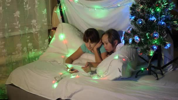 Sœurs avec tablette jouent dans une salle décorée avec arbre de Noël coloré. concept d'un Noël familial. enfants le soir de Noël jouer sur tablette, dans la chambre des enfants sur guirlandes canapé. — Video