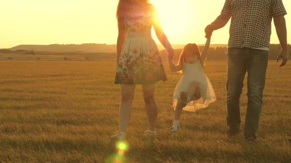 Mała córka skoki trzymając ręce taty i mama w parku na tle słońca. Koncepcja rodzinna. dziecko bawi się z ojcem i mamą na polu w świetle zachodu słońca. Chodzenie z małym dzieckiem w przyrodzie. — Zdjęcie stockowe
