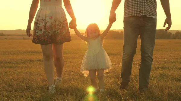 Liten dotter hoppar hålla händerna på pappa och mamma i parken på bakgrund av solen. Familjekoncept. barn leker med pappa och mamma på fältet i solnedgången ljus. Promenader med en liten unge i naturen. — Stockfoto