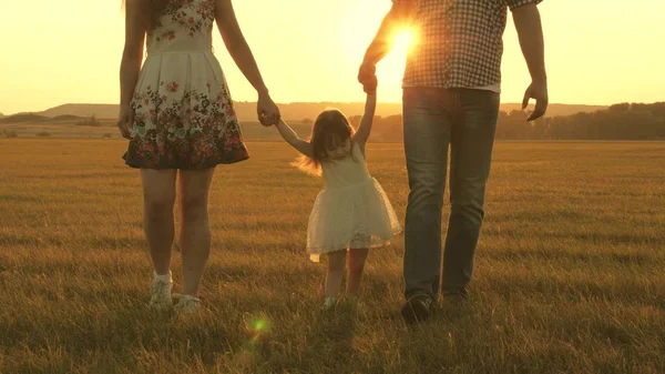 Μικρή κόρη πηδάει κρατώντας τα χέρια του μπαμπά και της μαμάς στο πάρκο στο φόντο του ήλιου. Οικογενειακή ιδέα. παιδί παίζει με τον μπαμπά και τη μαμά στο γήπεδο στο φως του ηλιοβασιλέματος. Περπάτημα με ένα μικρό παιδί στη φύση. — Φωτογραφία Αρχείου