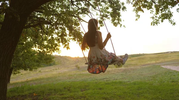 Barn som svänger på en gunga i parken i solen. ung flicka svänger på rep gunga på en ek gren. tonåring flicka njuter av flygning på swing på sommaren kväll i skogen. begreppet lycklig familj och barndom. — Stockfoto