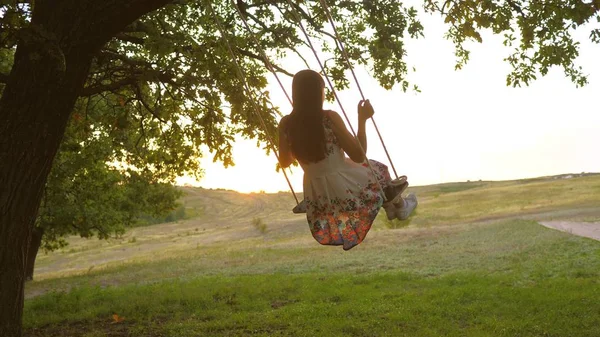 Niño balanceándose en un columpio en el parque bajo el sol. joven chica balanceándose en cuerda columpio en una rama de roble. adolescente chica disfruta de vuelo en swing en verano por la noche en el bosque. concepto de familia feliz y la infancia. — Foto de Stock