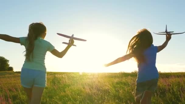 Uçma hayalleri. Mutlu çocukluk kavramı. Gün batımında iki kız bir oyuncak uçakla oynuyor. Güneşin arka planındaki çocuklar ellerinde bir uçakla. Uçakta oynayan çocukların silueti — Stok video