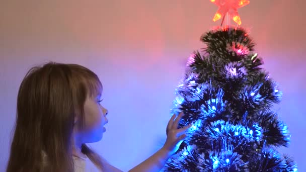 La niña juega junto al brillante árbol de navidad en el vivero. niño examina guirnalda en el árbol de Navidad. hermoso árbol de Navidad artificial. concepto de infancia feliz. niño juega para las vacaciones de Navidad — Vídeo de stock